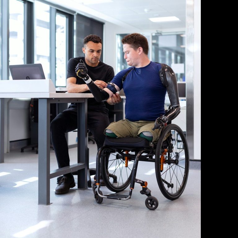 Ein Mann im Rollstuhl mit Prothese sitzt am Tisch, während ein anderer ihn berät.